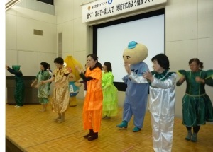 舞台上でカラフルな衣装を着て立っている出演者とたじりっちとトライ君の写真