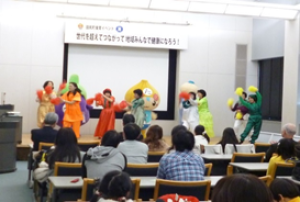 舞台上でたじりっちや、なすびんと一緒に踊っている出演者と席に座って見ている食育講演会の参加者の写真