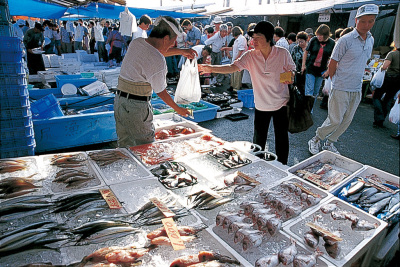 日曜朝市で、店先には沢山の新鮮な魚が並んでおり、買い物に来たい女性客が店主から購入した品物を手渡しで受け取っている写真