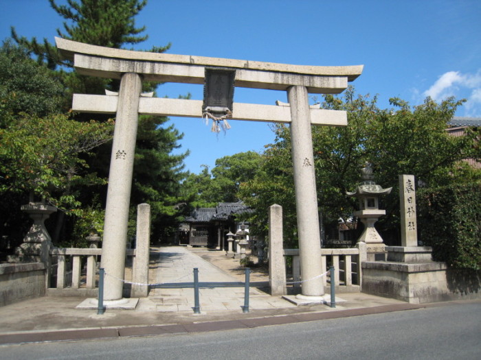 青空の下、鳥居を真ん中にし、両端に緑の樹木が続く春日神社入り口を写した写真