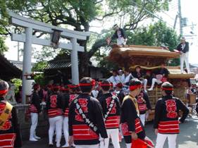 鳥居の横に神輿が運ばれ、赤いねじり鉢巻きに法被を着た沢山の方々が祭りに参加している様子の写真