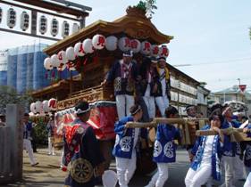提灯が沢山下げられた大きな神輿に3人の男性が乗っており、青い法被を着た参加者の方々が神輿を力いっぱい動かしている秋祭りの様子の写真