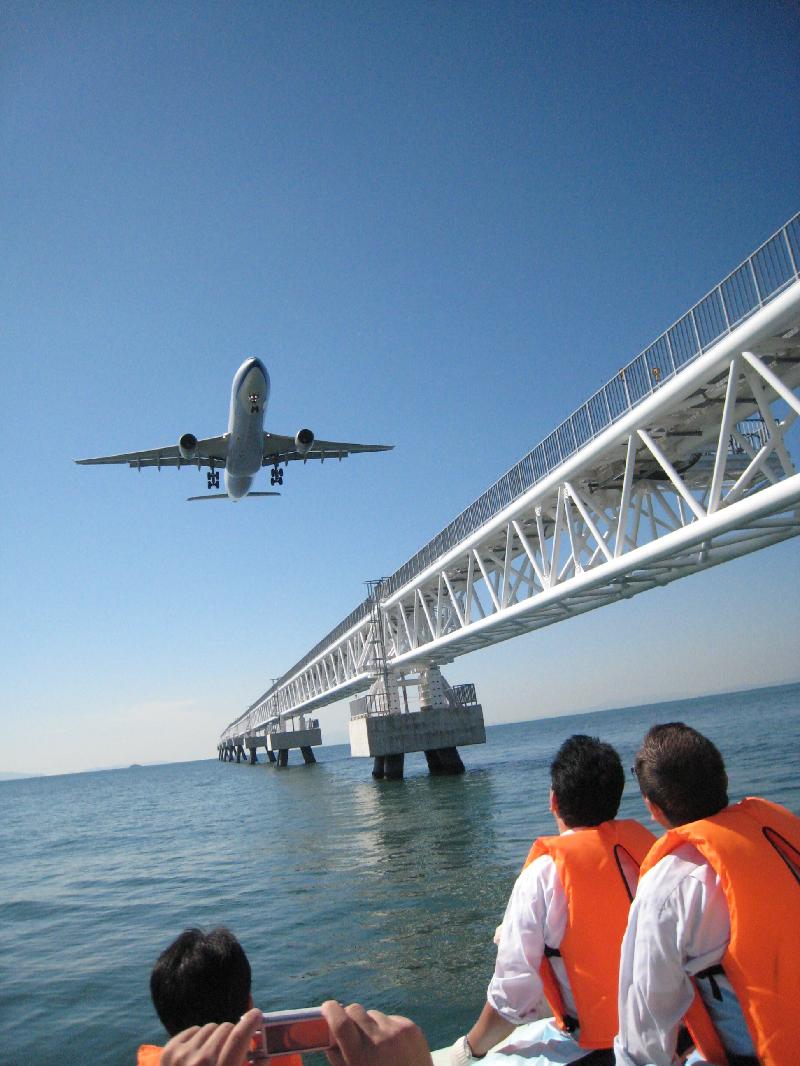 海の上に真っすぐに渡っている大きな橋のすぐ横を飛んでいる飛行機とその様子を海上から見上げている関空クルーズの方々の写真