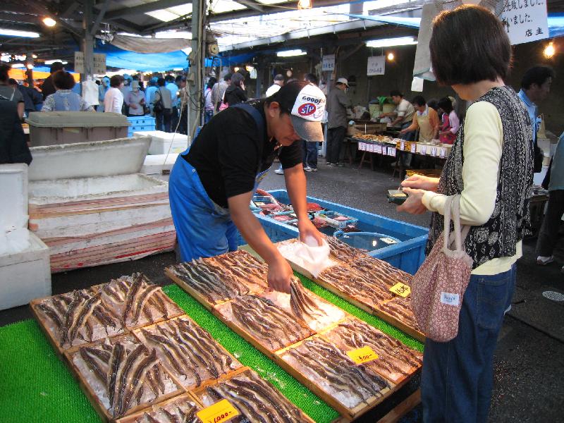 店先にたくさんの魚が並んでいて、朝市に買い物に来た女性が魚を購入している田尻漁港日曜朝市の賑わいを見せている様子を写した写真