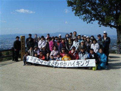 青空の下平成24年度さわやかハイキングに参加した人達が横断幕を持ち記念撮影をしている写真