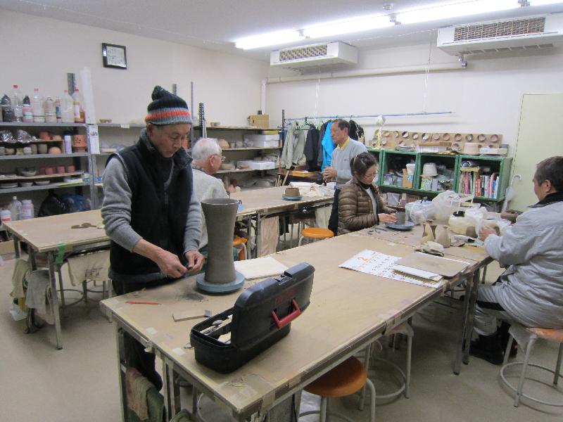 男性が机の上で粘土で陶芸作品を作成している奥では他の人達が椅子に座り作品造りをしている写真