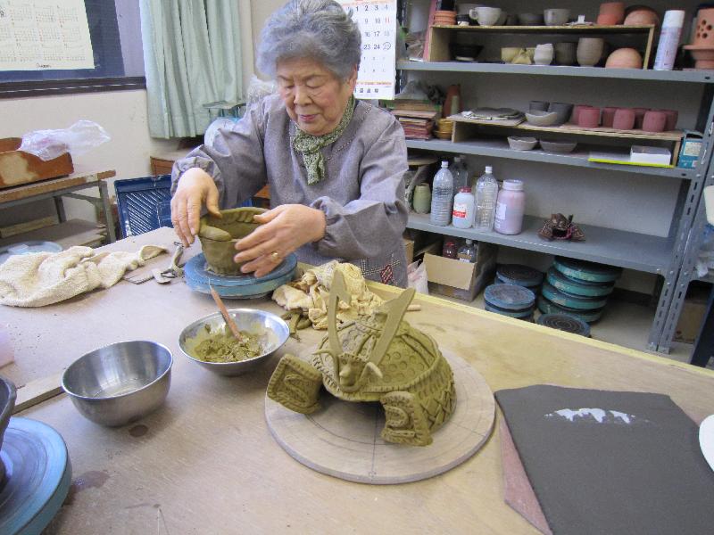 女性が粘土をこねている手前には粘土で造られた兜が置かれている写真