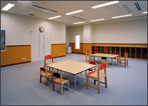 絨毯が敷かれた室内で、中央に間隔を空けて2つの机が置かれ、木製の子ども用の椅子と大人用の椅子が設置されているプレイルームの写真