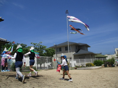 風にたなびいているこいのぼりと手作りのこいのぼりを持って園庭を走っている園児たちの写真