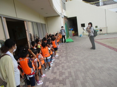 ライフジャケットを着て並んで手を挙げている園児たちと話をしている園長先生の写真