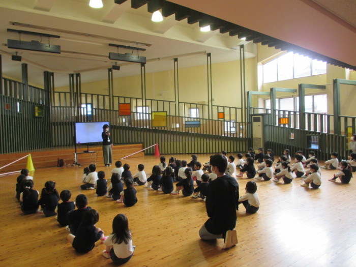 前方に立って話をしている先生と整列して床に座り話を聞いている園児たちを後ろから写した写真