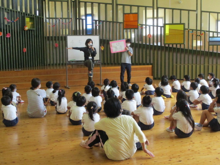 右手をまっすぐ横に伸ばして右足を挙げて片足で立って話をしている先生と床に座って話を聞いている園児たちの写真