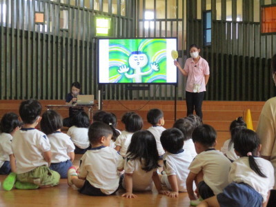 前方モニター横に立って話をしている先生と床に座って話を聞いている園児たちの写真