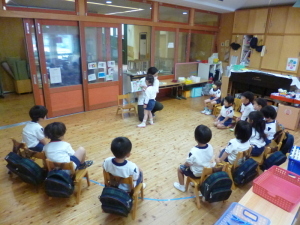 絵本を読み聞かせしている先生と立ち上がって絵本を見ている園児、椅子に座っている園児たちの写真