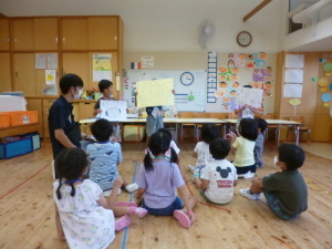 紙を持って前に立っている園児と座って聞いている園児たちと先生の写真