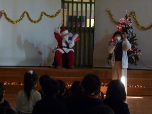 クリスマスツリーの横の椅子に腰かけているサンタクロースとマイクを持って話をしている先生の写真