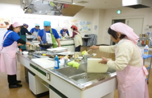 調理室で鍋をかき混ぜたりまな板を洗ったりしている調理実習参加者の写真