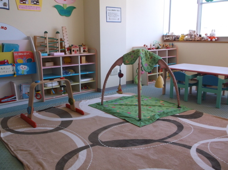 壁際に本棚や遊具を片付ける棚が設置され、子ども用の机と椅子、部屋の中央には絨毯が敷かれ、アーチにおもちゃが吊り下げられている赤ちゃん用のメリージムが置かれているすくすくセンターの室内写真